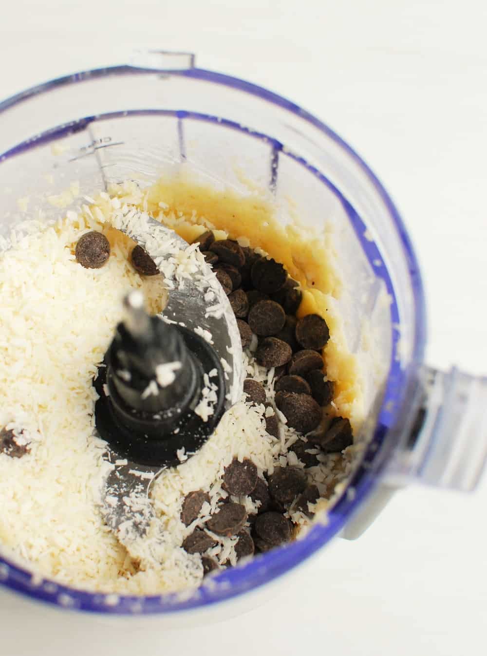 a food processor bowl with processed macadamia nuts, and then chocolate chips and coconut added on top