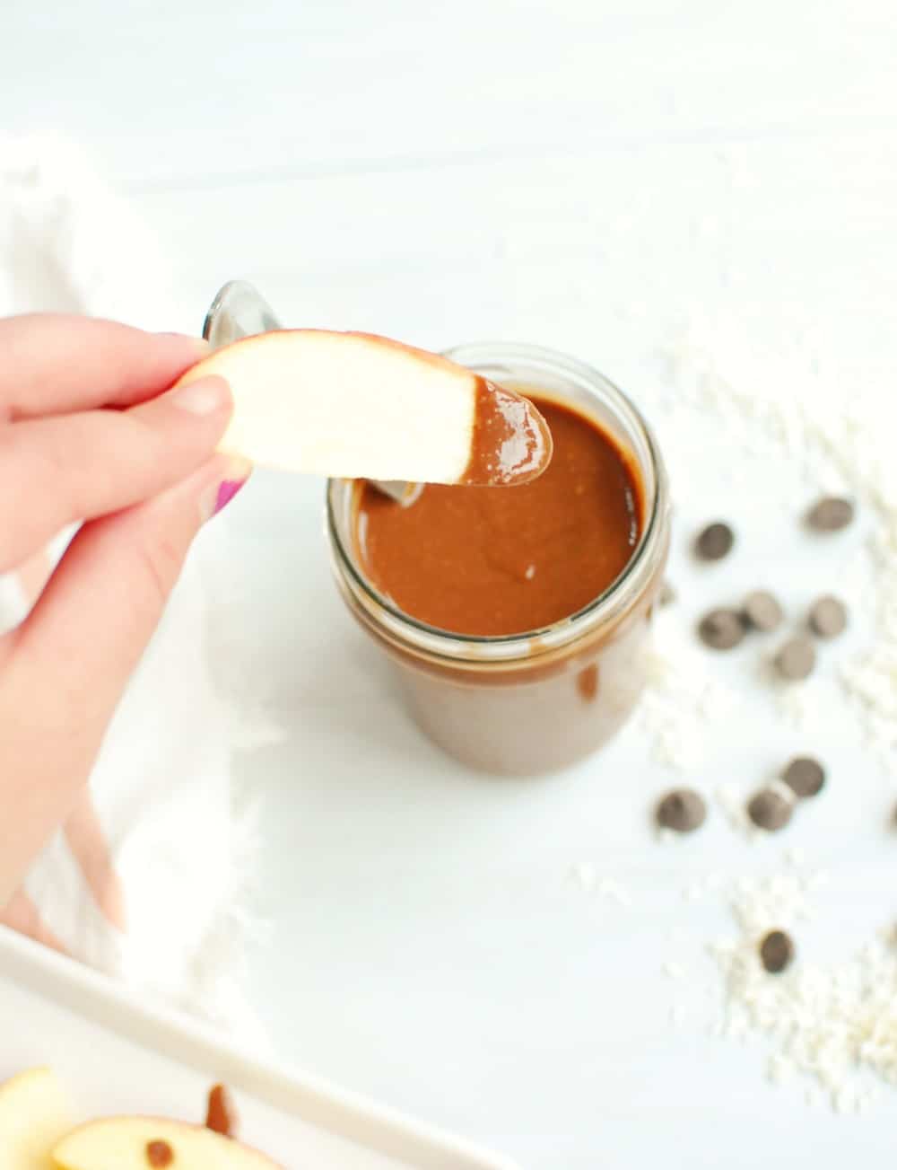 a woman dipping a slice of apple into a jar of chocolate macadamia nut butter