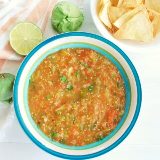 a bowl full of tomatillo and tomato salsa, next to a lime, some tomatillos, and some tortilla chips