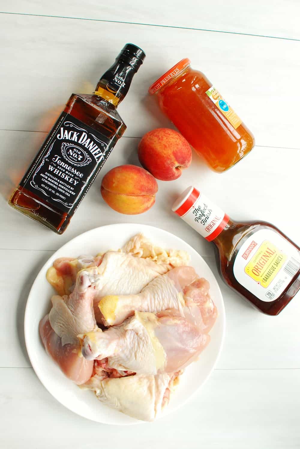 a plate of chicken thighs and drumsticks, a bottle of barbecue sauce, two peaches, whiskey, and a jar of apricot preserves