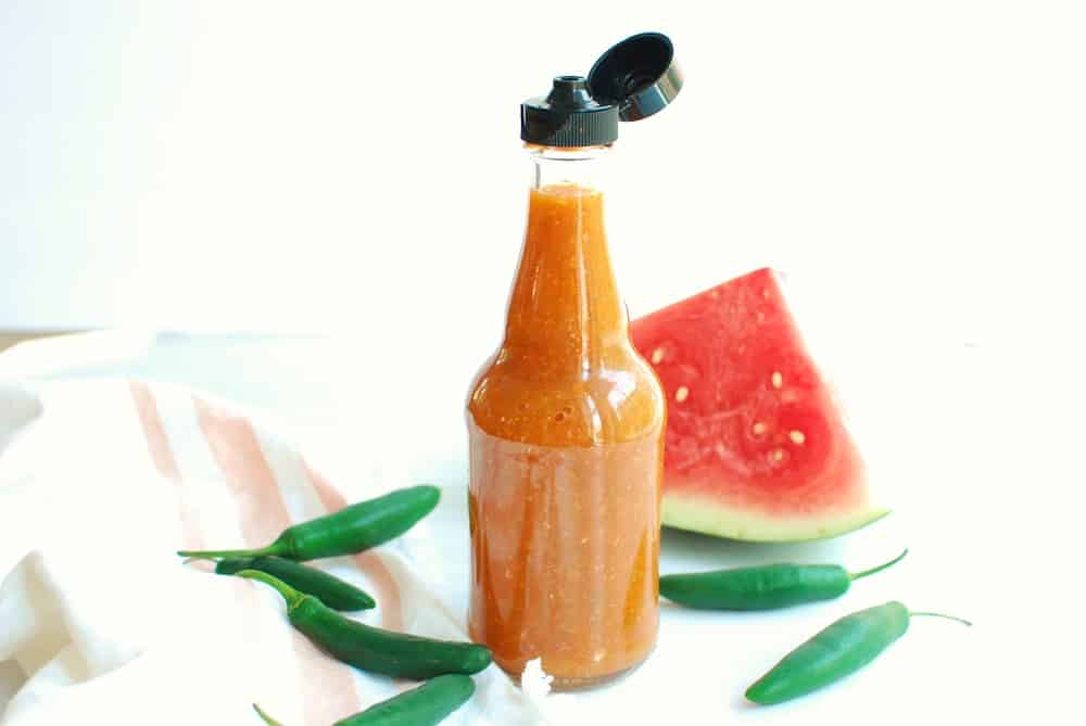 a bottle of homemade watermelon hot sauce next to a napkin and some serrano peppers