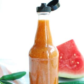 a bottle of watermelon hot sauce next to some serrano peppers and fresh watermelon