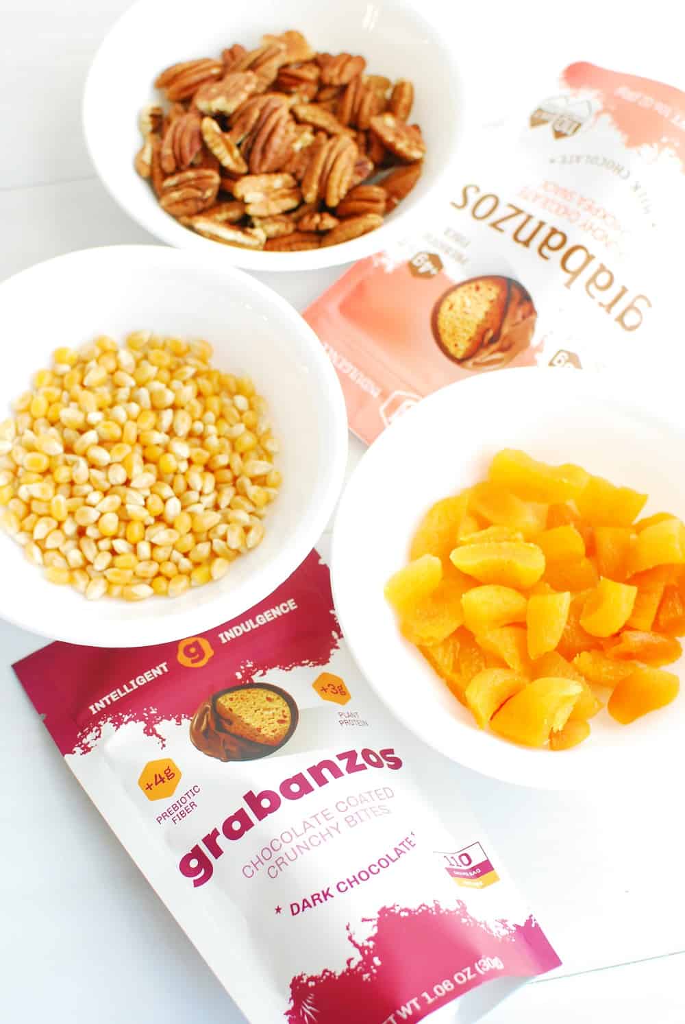 a bag of grabanzos, a bowl of popcorn kernels, a bowl of dried apricots, and a bowl of pecans