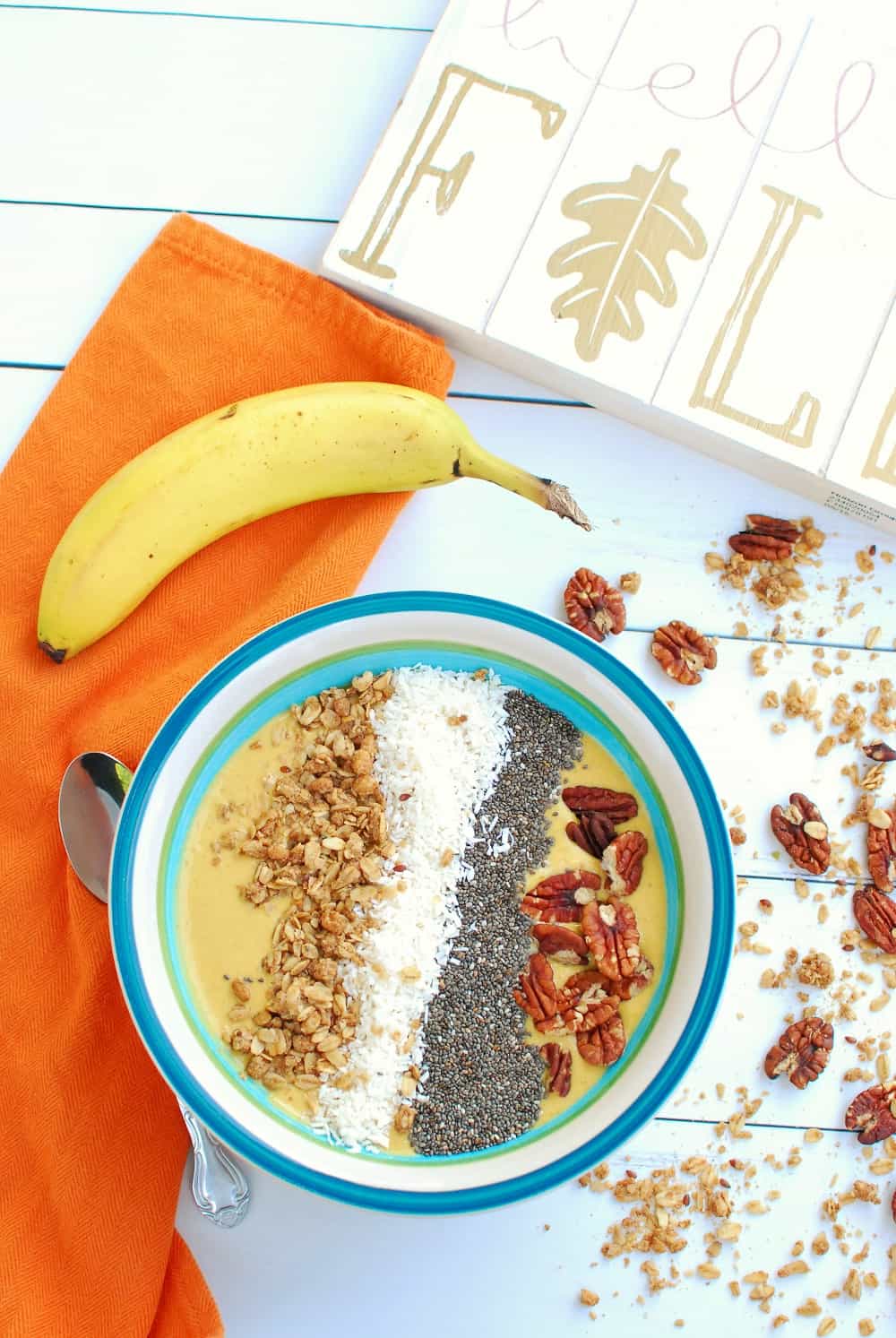 a pumpkin smoothie bowl next to an orange napkin, a banana, and some scattered granola and pecans