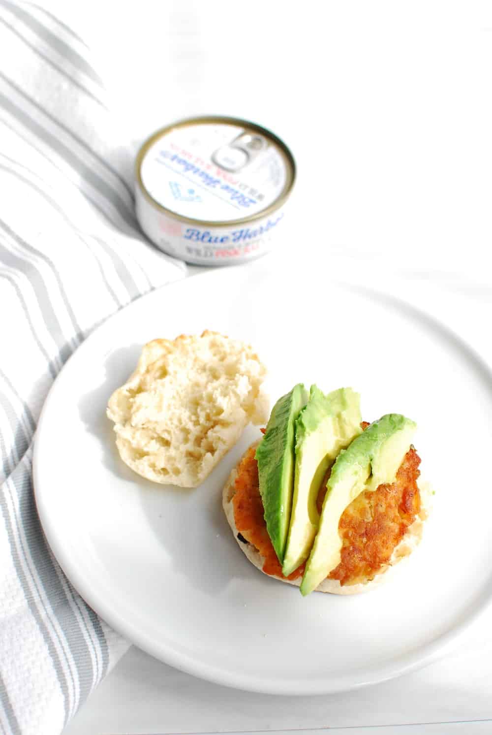 an english muffin topped with a salmon patty and several slices of avocado, next to a can of salmon