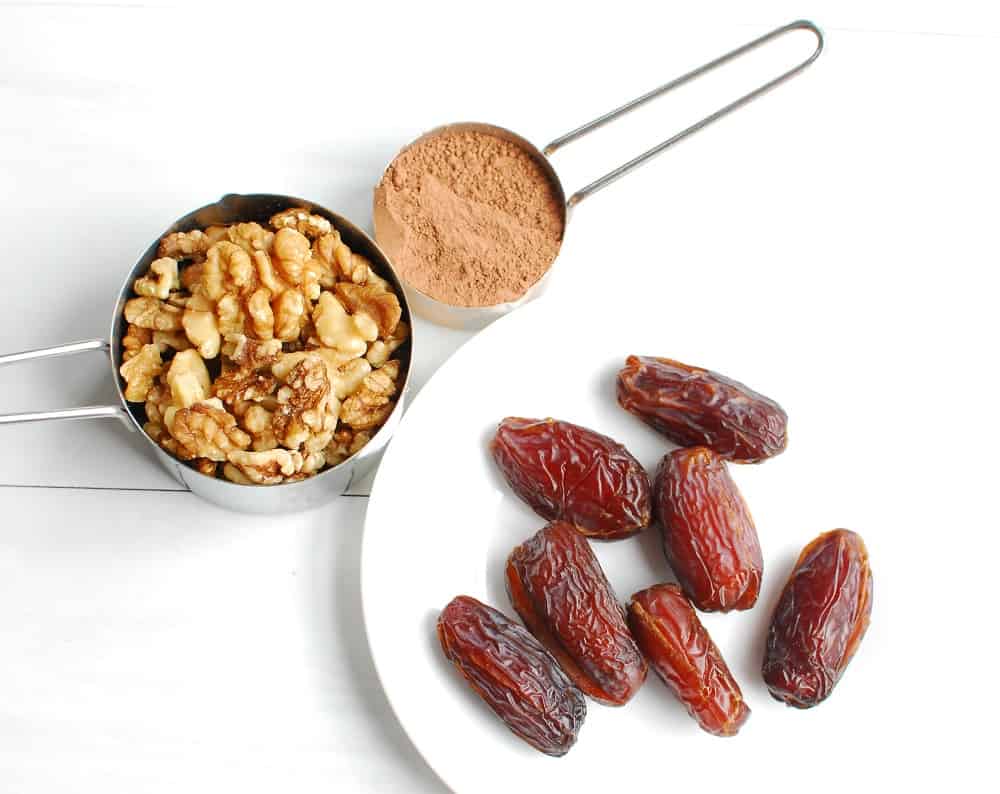 dates on a plate, walnuts in a measuring cup, and cocoa powder in a measuring cup