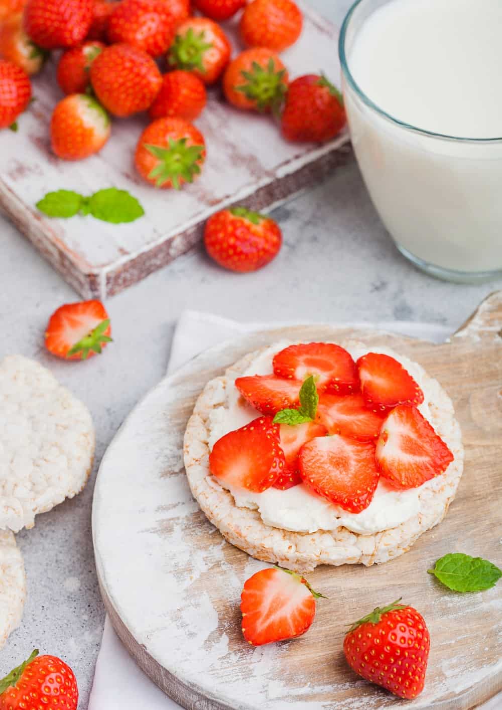 Healthy organic rice cakes with ricotta and fresh strawberries on wooden board and glass of milk on light stone kitchen background.