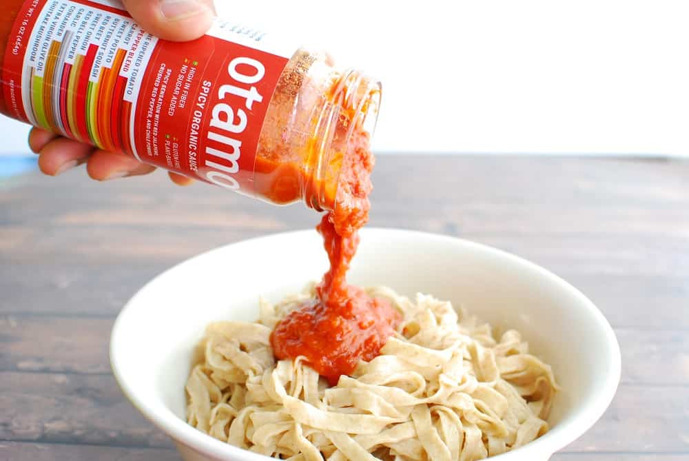 a man's hand pouring otamot tomato sauce onto a bowl of cooked pasta noodles