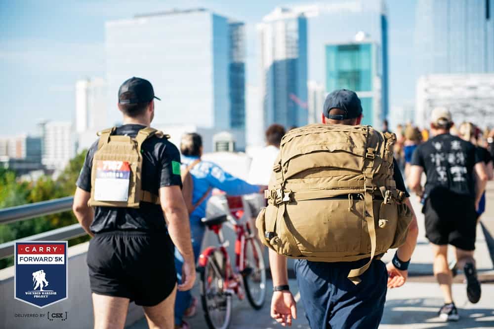 two men carrying weighted backpacks walking in a 5K race