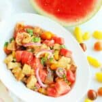 a bowl full of watermelon panzanella salad next to some cherry tomatoes and a slice of watermelon