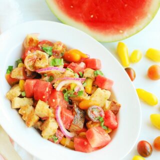 a bowl full of watermelon panzanella salad next to some cherry tomatoes and a slice of watermelon