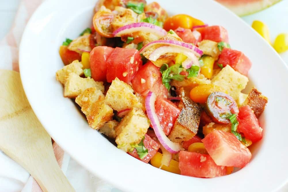 a white serving bowl filled with watermelon panzanella salad next to a wooden spoon