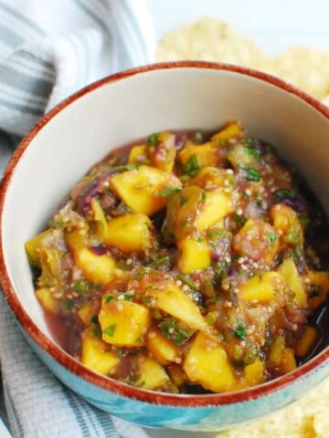 a bowl of mango tomatillo salsa next to a spoon, napkin, and chips