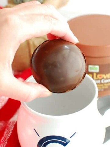 A woman holding a hot chocolate bomb about to put it in a coffee mug.