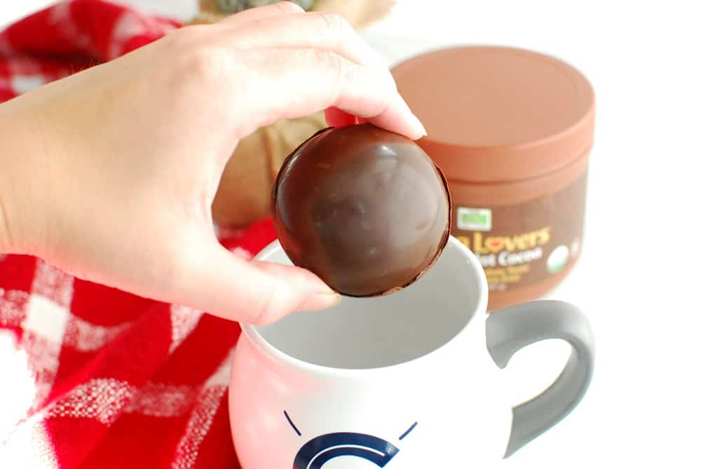 A woman holding a hot chocolate bomb about to put it in a coffee mug.