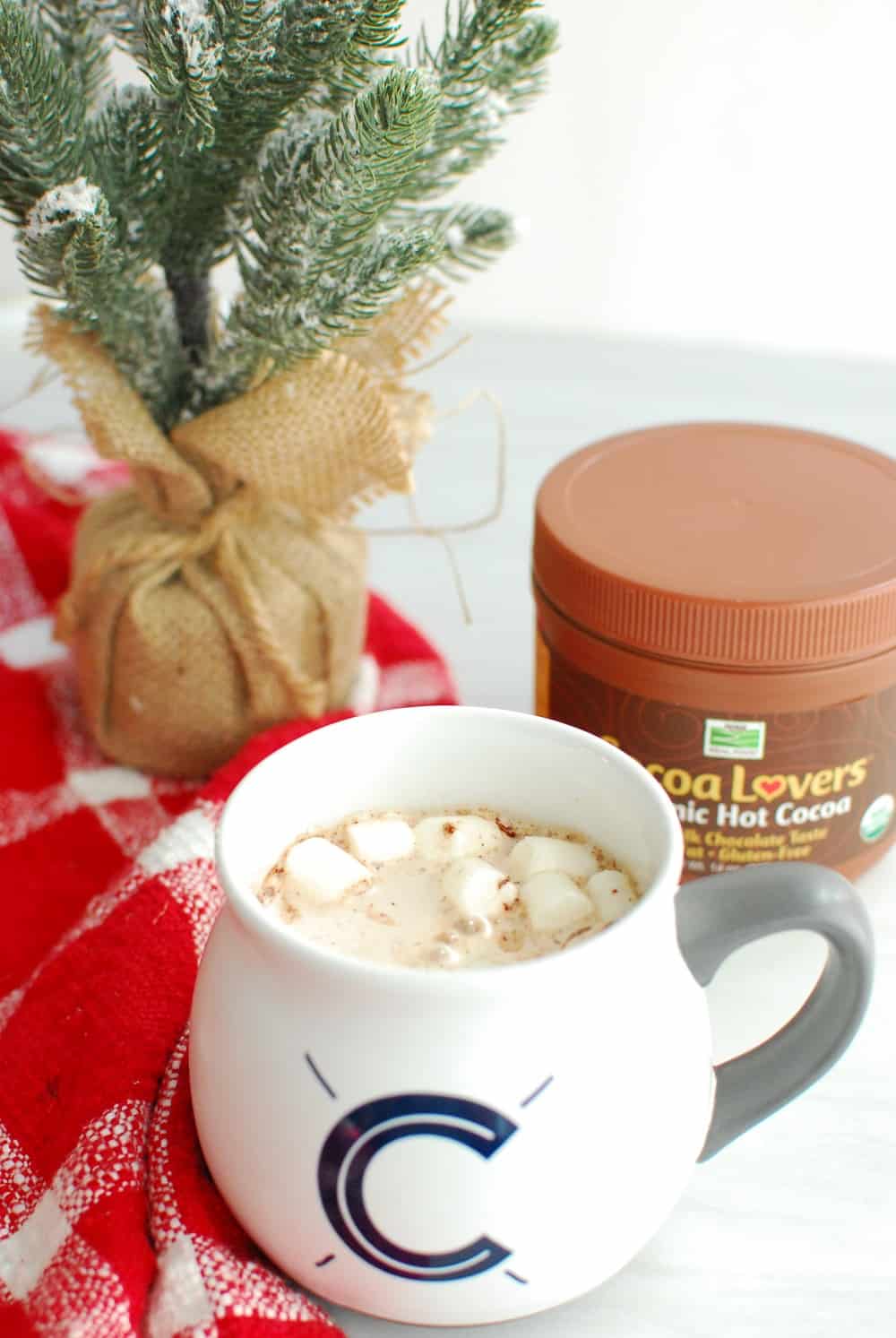 a cup of hot chocolate with marshmallows, next to a mini christmas tree and a container of hot chocolate