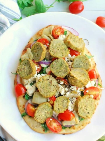 a Mediterranean flatbread on a plate next to a fork and knife, as well as some loose arugula and cherry tomatoes