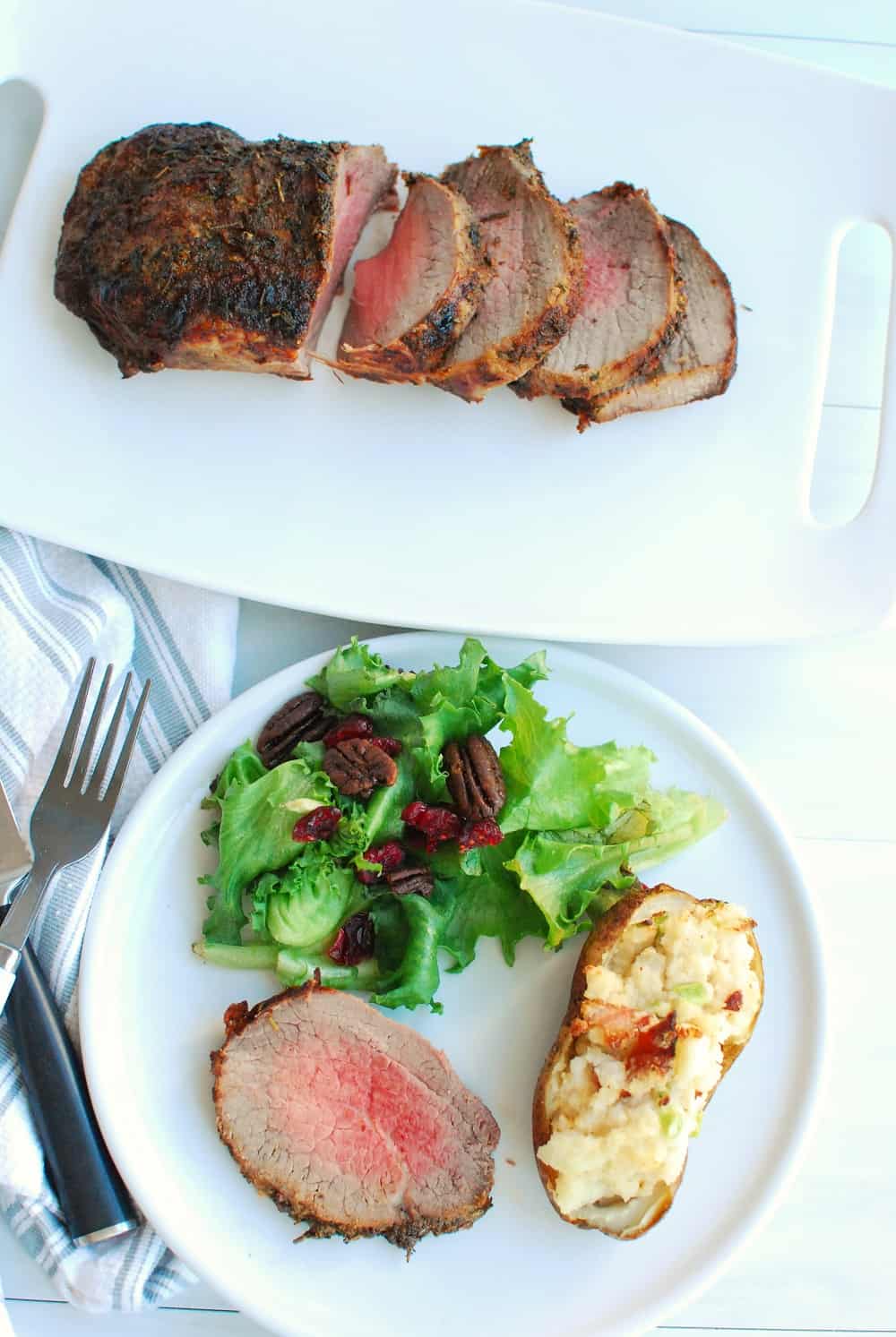 an eye of round roast on a plate with salad and a twice baked potato, next to a platter with the rest of the roast