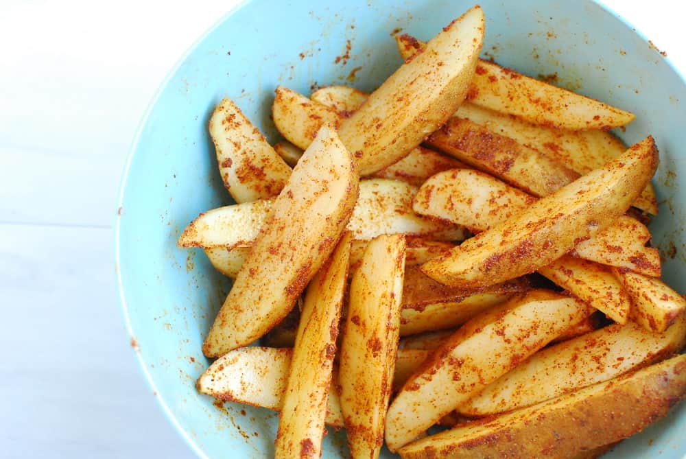 Potato wedges tossed in oil and spices in a blue mixing bowl.