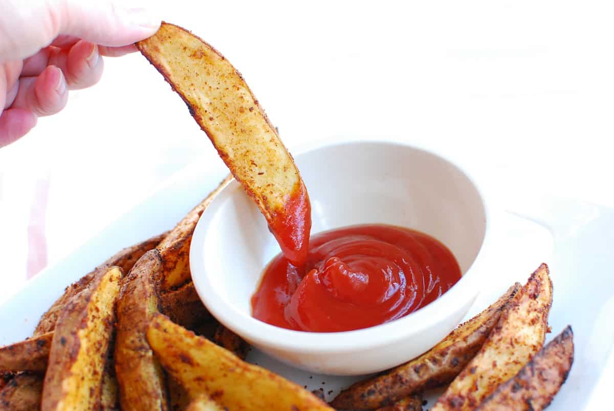 A woman's hand holding a potato wedge and dipping it in ketchup.