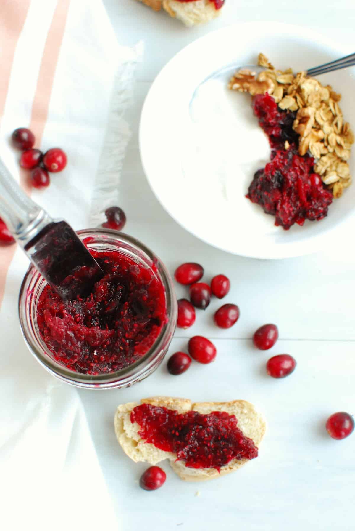 A jar of cranberry chia jam next to bread with the jam spread on it and a bowl of yogurt with the jam mixed in.