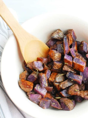 A bowl of herb roasted purple potatoes with a wooden spoon.