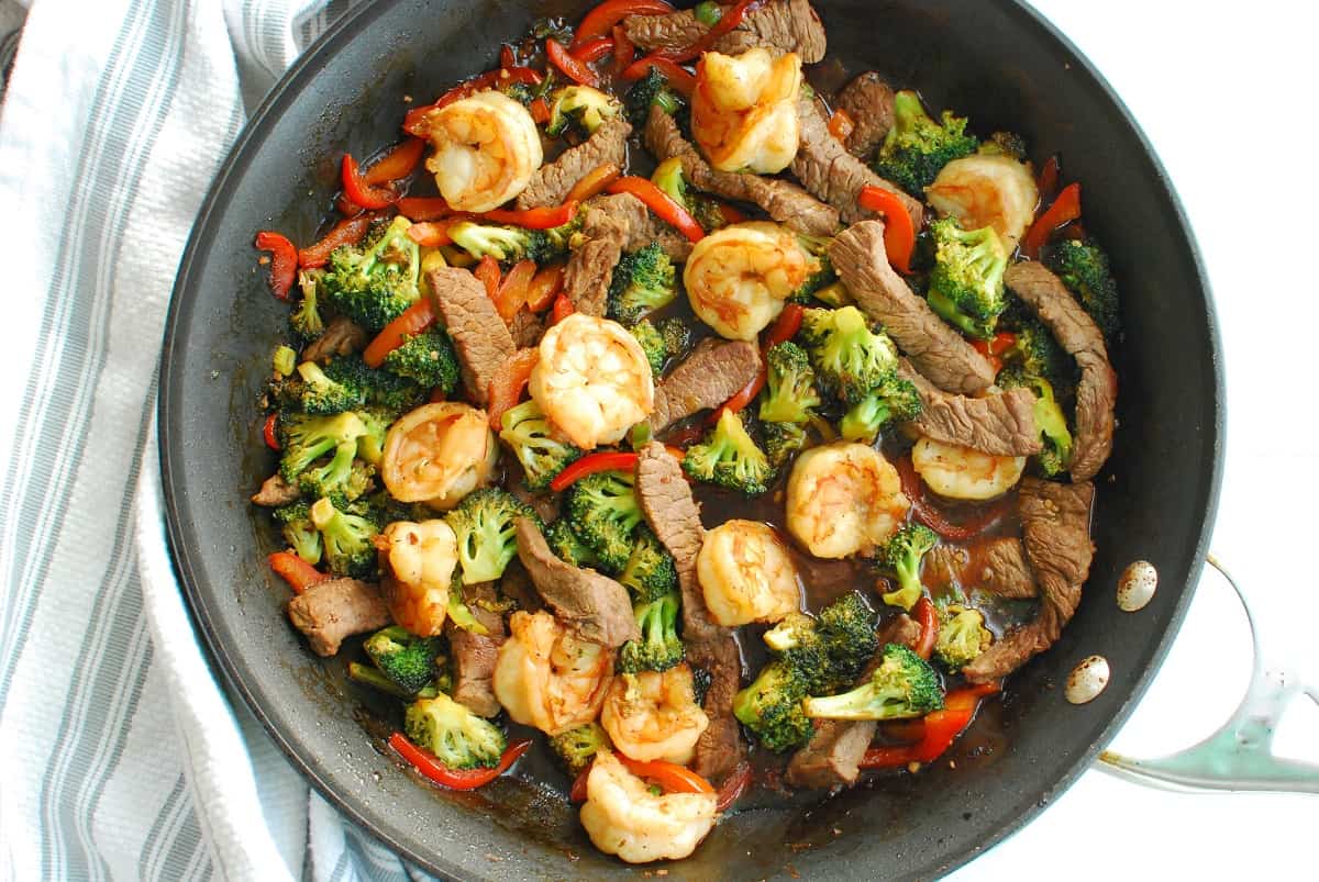 A skillet of shrimp and steak stir fry next to a striped napkin.