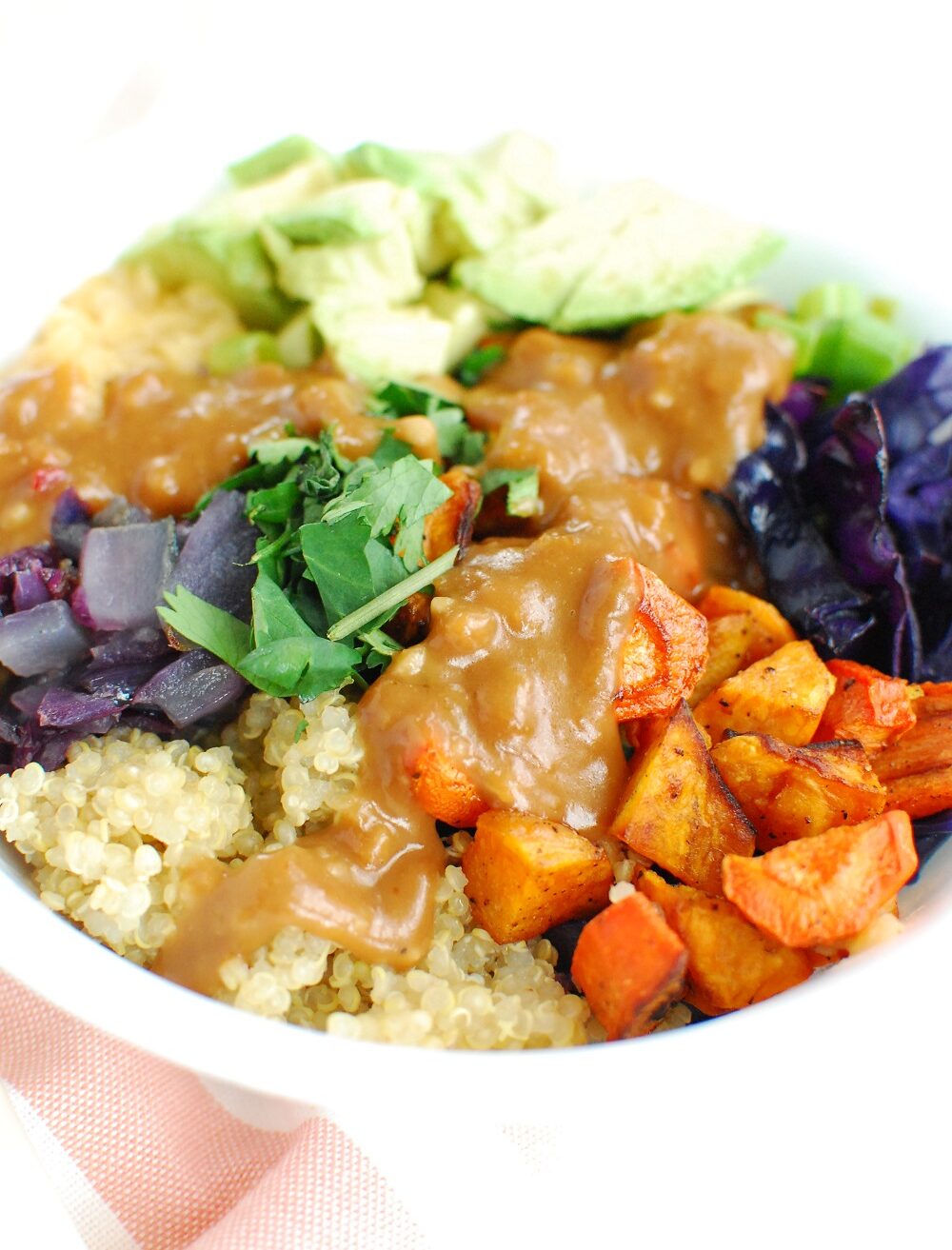 A white bowl filled with quinoa, lentils, carrots, sweet potatoes, cilantro, cabbage, onion, and avocado, topped with peanut sauce.