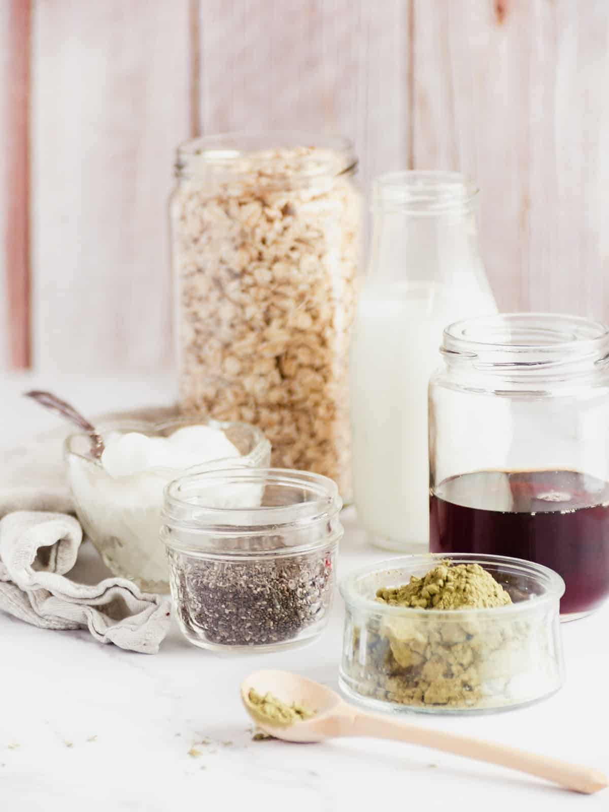 A bowl of matcha, a jar of rolled oats, a jar of milk, a jar of maple syrup, a jar of chia seeds, and a bowl of yogurt.