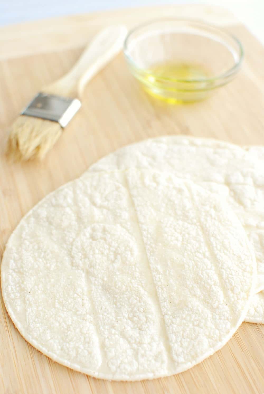 Corn tortillas on a cutting board next to olive oil and a brush.