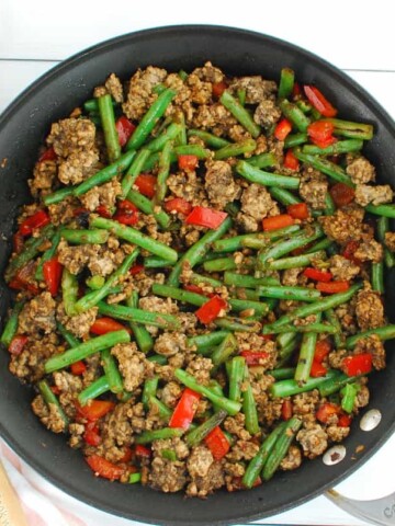 Ground pork stir fry with green beans in a large skillet next to a wooden spoon.
