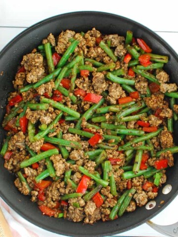 Ground pork stir fry with green beans in a large skillet next to a wooden spoon.