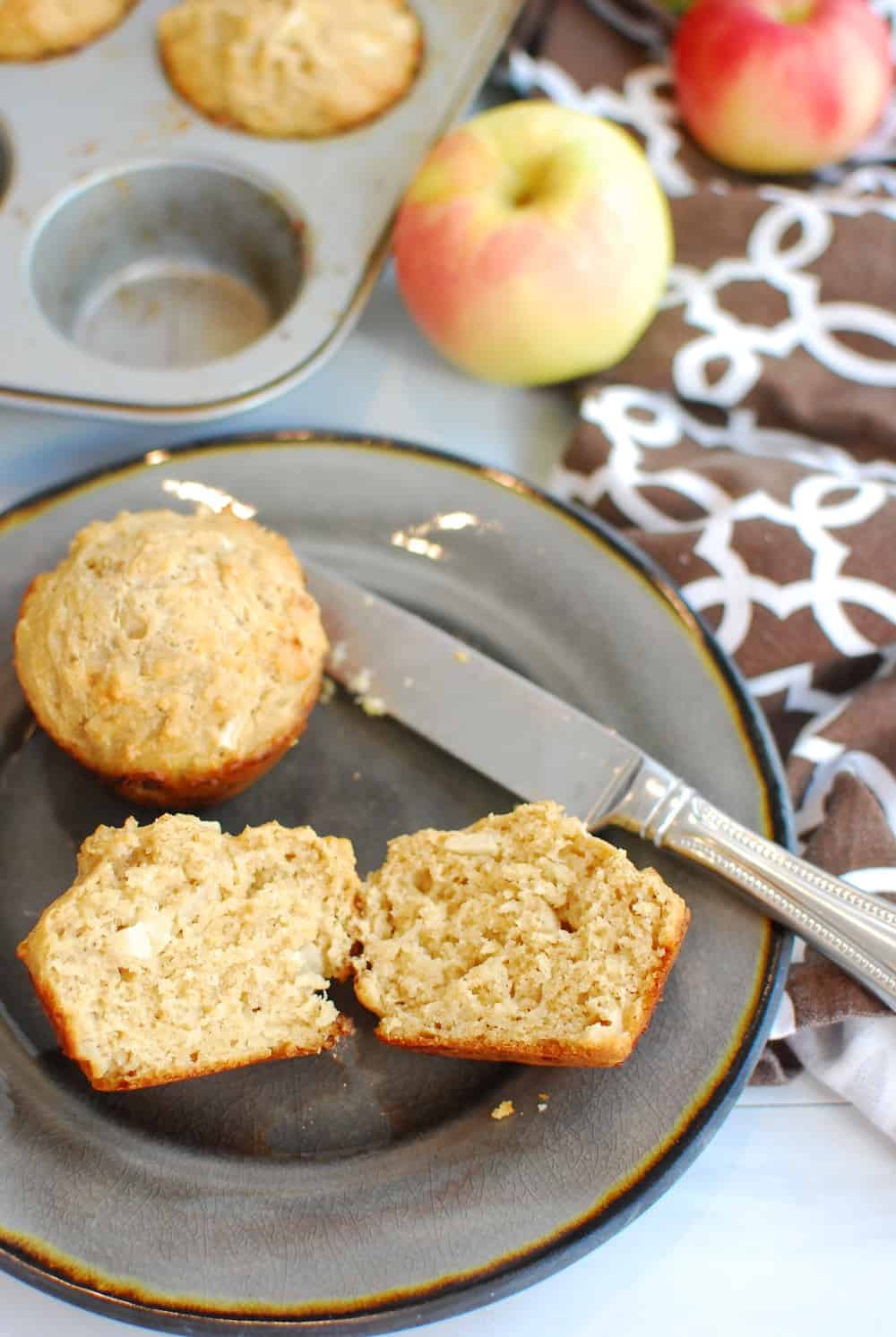 An apple cheddar muffin on a plate that was cut in half, next to a napkin and an apple.