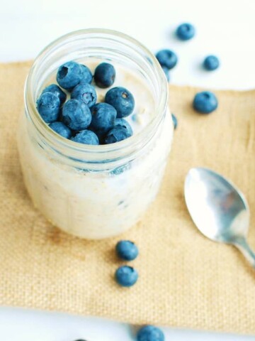 A mason jar with high protein overnight oats sitting on rustic cloth next to a spoon and some scattered blueberries.