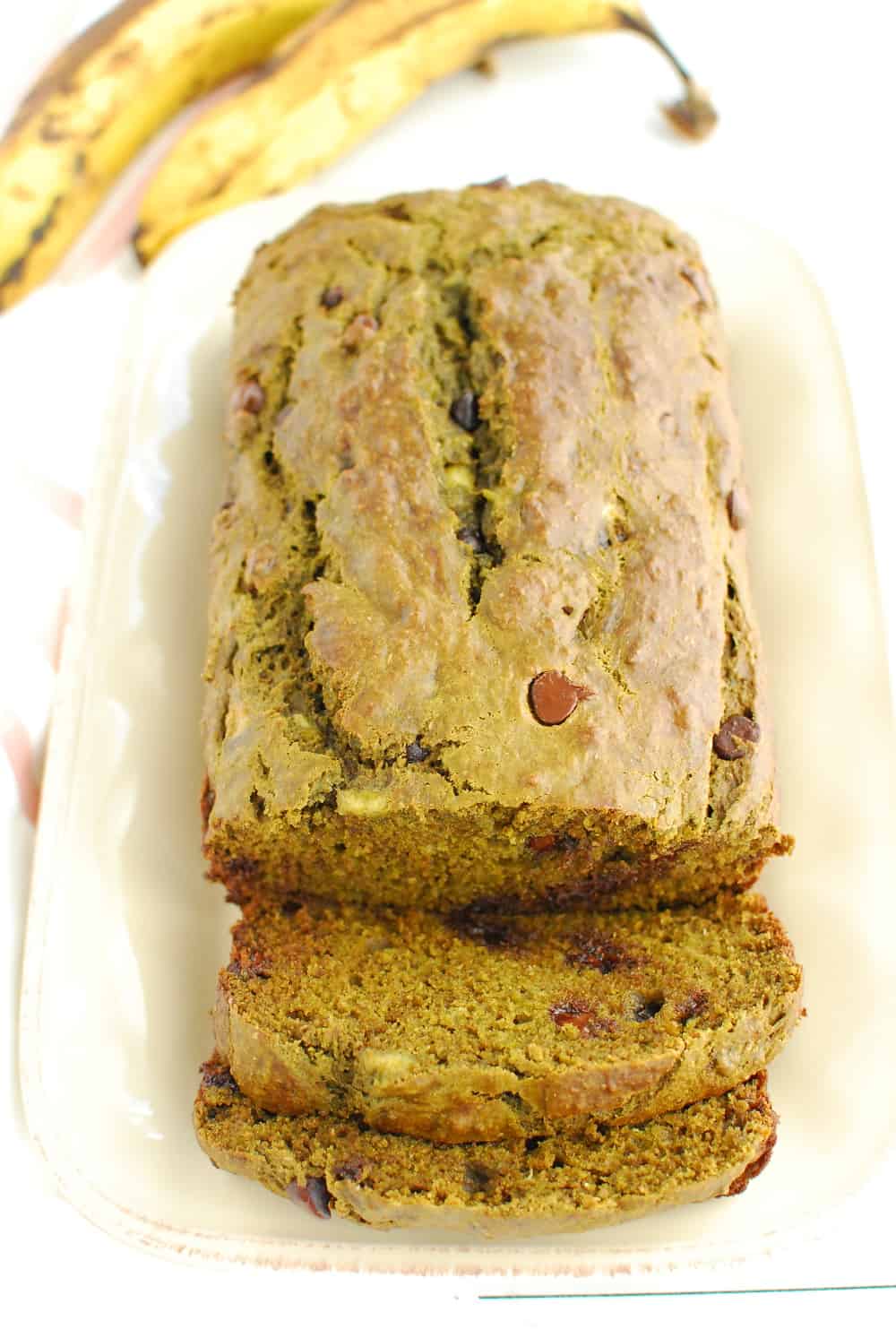 A loaf of matcha banana bread that has been sliced into, next to two overripe bananas.