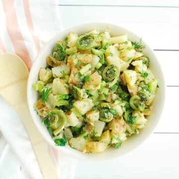 A bowl of vegan dill potato salad with fiddleheads next to a striped napkin and a wooden serving spoon.