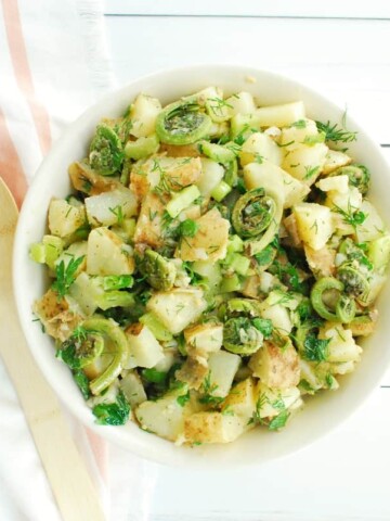 A bowl of vegan dill potato salad with fiddleheads next to a striped napkin and a wooden serving spoon.