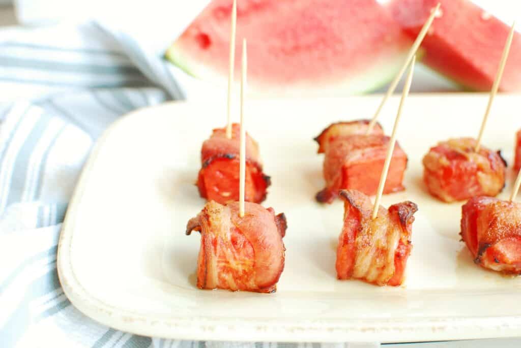 Several pieces of bacon wrapped watermelon on a plate next to a striped napkin.