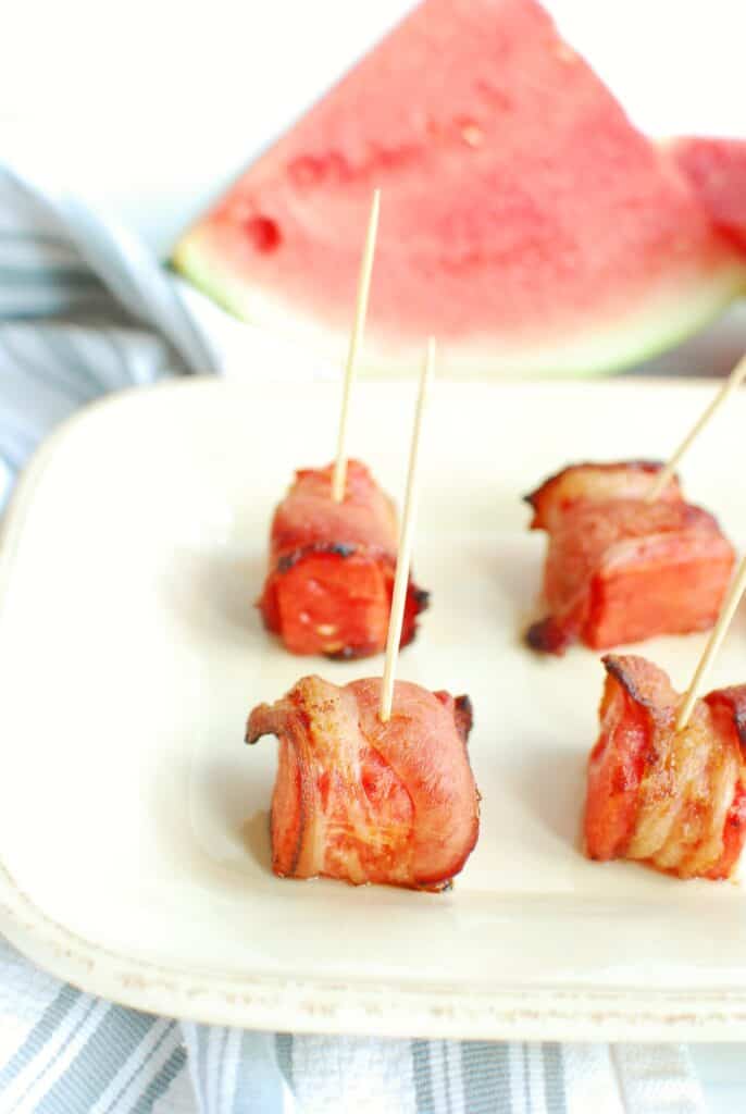 Several pieces of bacon wrapped watermelon on a plate, with a slice of watermelon in the background.