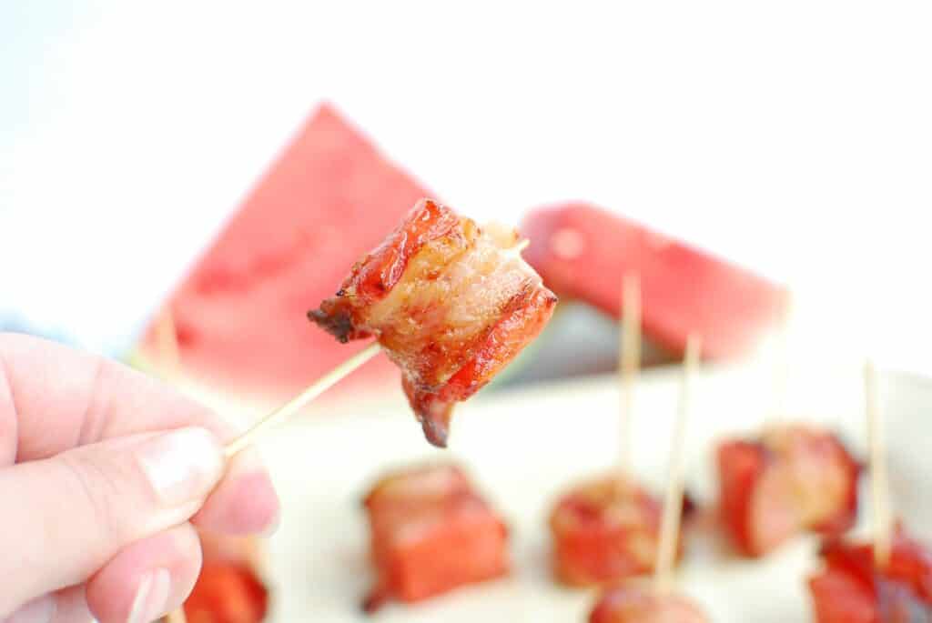 A piece of bacon wrapped watermelon held up by a toothpick.