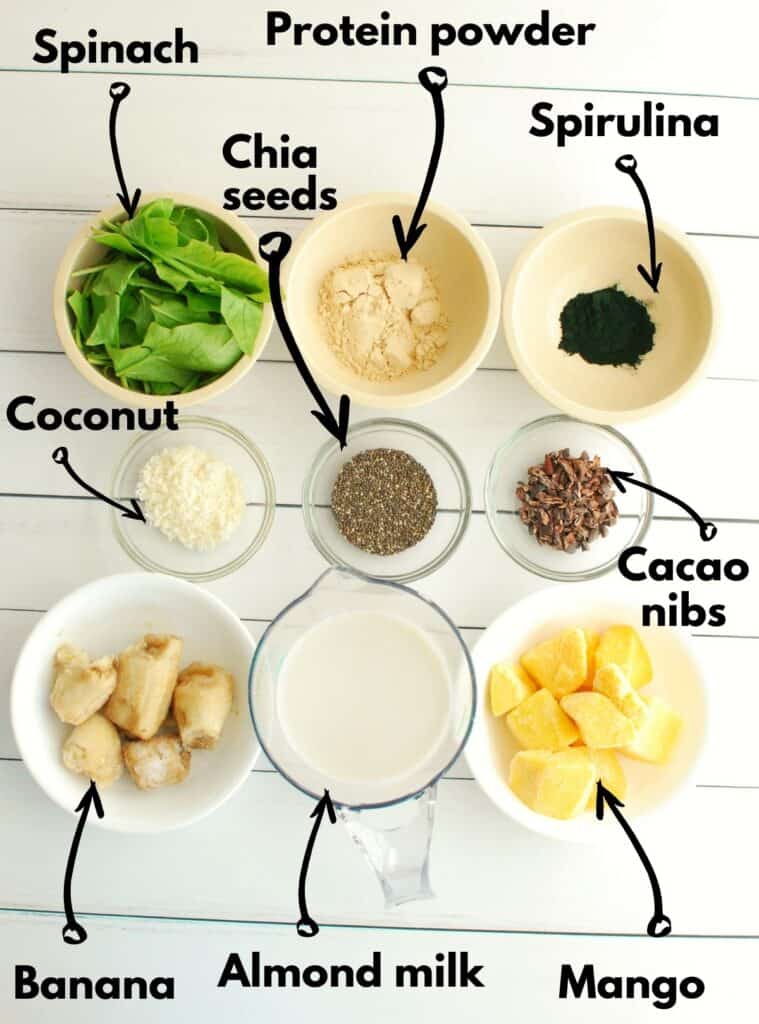 A shot of ingredients, including banana, almond milk, mango, spirulina, protein powder, spinach, chia seeds, coconut, and cacao nibs.