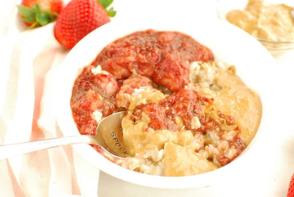 A spoon digging into a bowl of oatmeal, topped with peanut butter and strawberry chia jam.