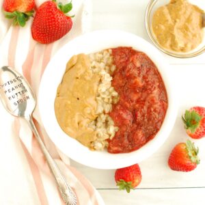 A bowl of peanut butter and jelly oatmeal next to a spoon, napkin, and fresh strawberries.