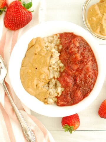 A bowl of peanut butter and jelly oatmeal next to a spoon, napkin, and fresh strawberries.