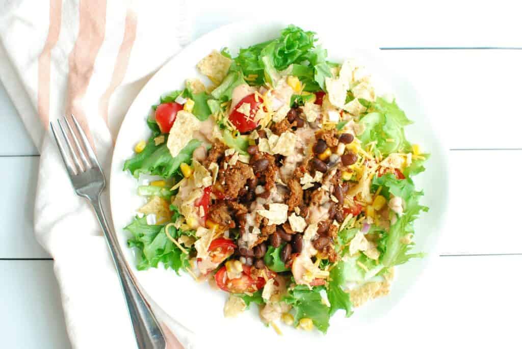 A taco salad on a white plate next to a fork and napkin.