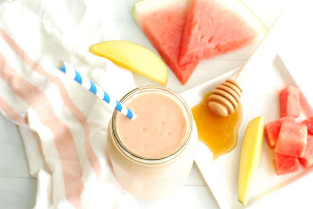 A watermelon protein shake in a mason jar next to some fresh watermelon, mango, and honey.