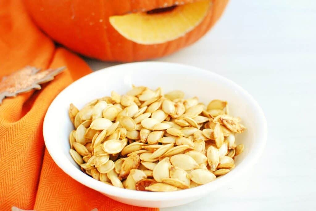 A close up of a bowl of pumpkin seeds next to a jack-o-lantern.