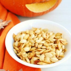 Air fryer roasted pumpkin seeds in a white bowl next to an orange napkin and a pumpkin.