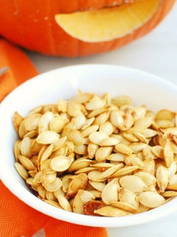 Air fryer roasted pumpkin seeds in a white bowl next to an orange napkin and a pumpkin.