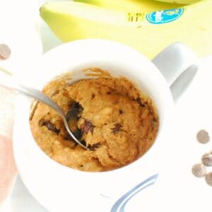 A spoon digging into a healthy banana mug cake, next to some fresh bananas and chocolate chips.